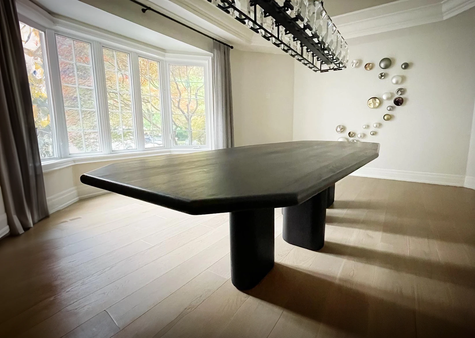 A modern dining room with an asymmetric dining table featuring cylindrical legs, a chandelier above, a bay window on the left, and decorative circular wall art on the right.