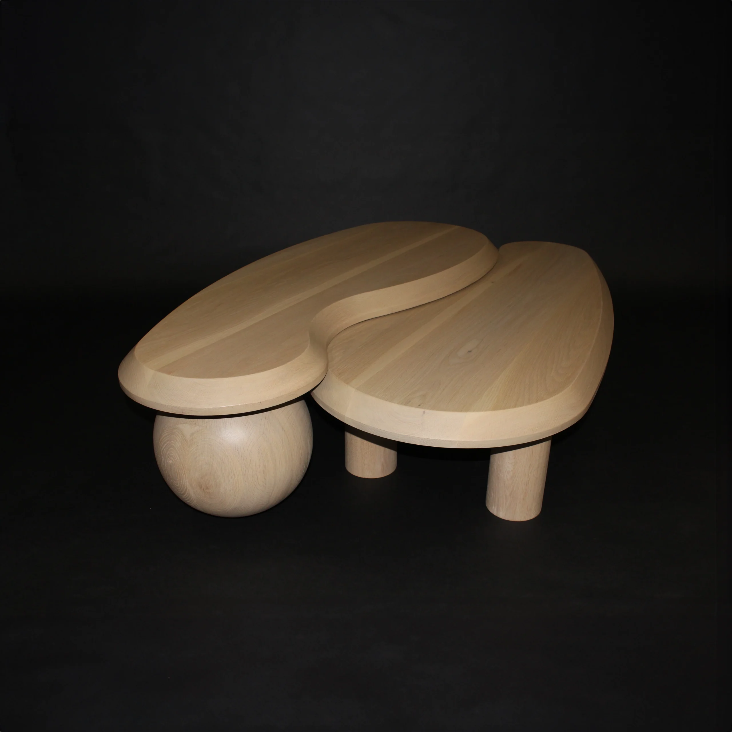 A modern luxury coffee table with an abstract design featuring two curved sections resting on cylindrical legs, and one section supported by a spherical base, set against a dark background.