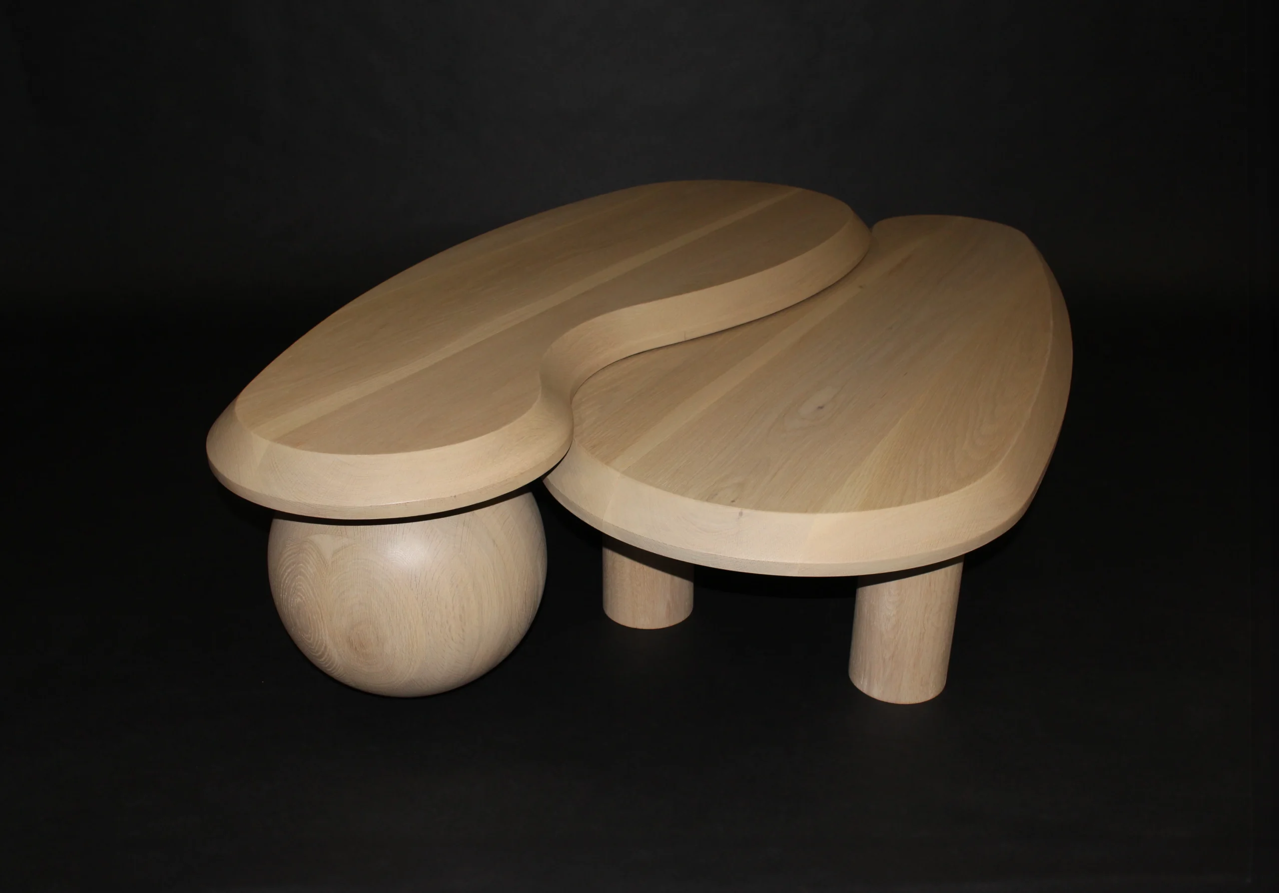 A wooden abstract sculpture featuring two curved, flat surfaces, one elevated on a spherical base and supported by cylindrical legs. This harmonious piece evokes the elegance of a modern luxury coffee table against a plain black background.