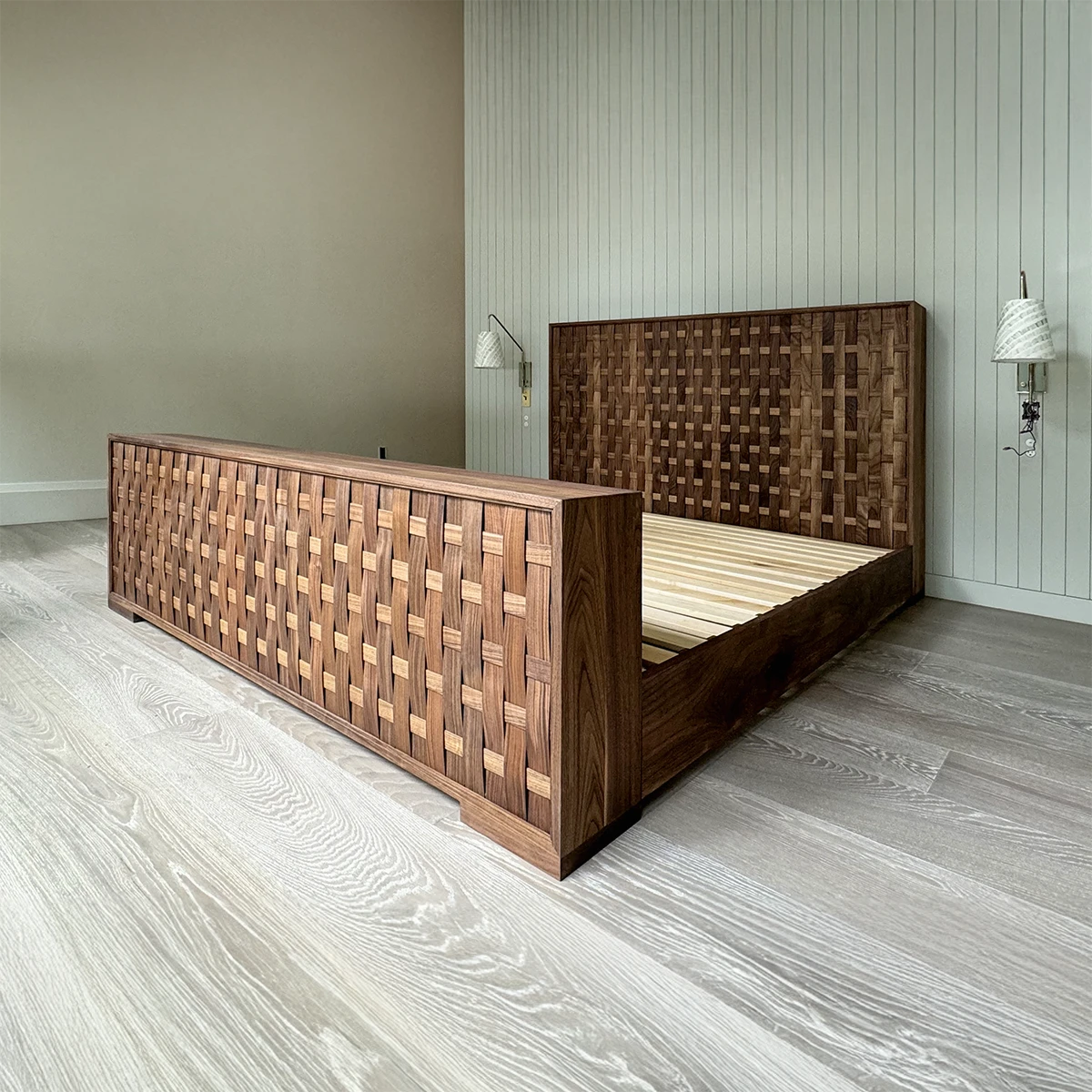 A luxury king size bed features a wooden frame with a woven pattern headboard and footboard, set against light-colored wood flooring and white paneled walls. Two wall-mounted lamps elegantly flank the bed, enhancing its sophisticated style.