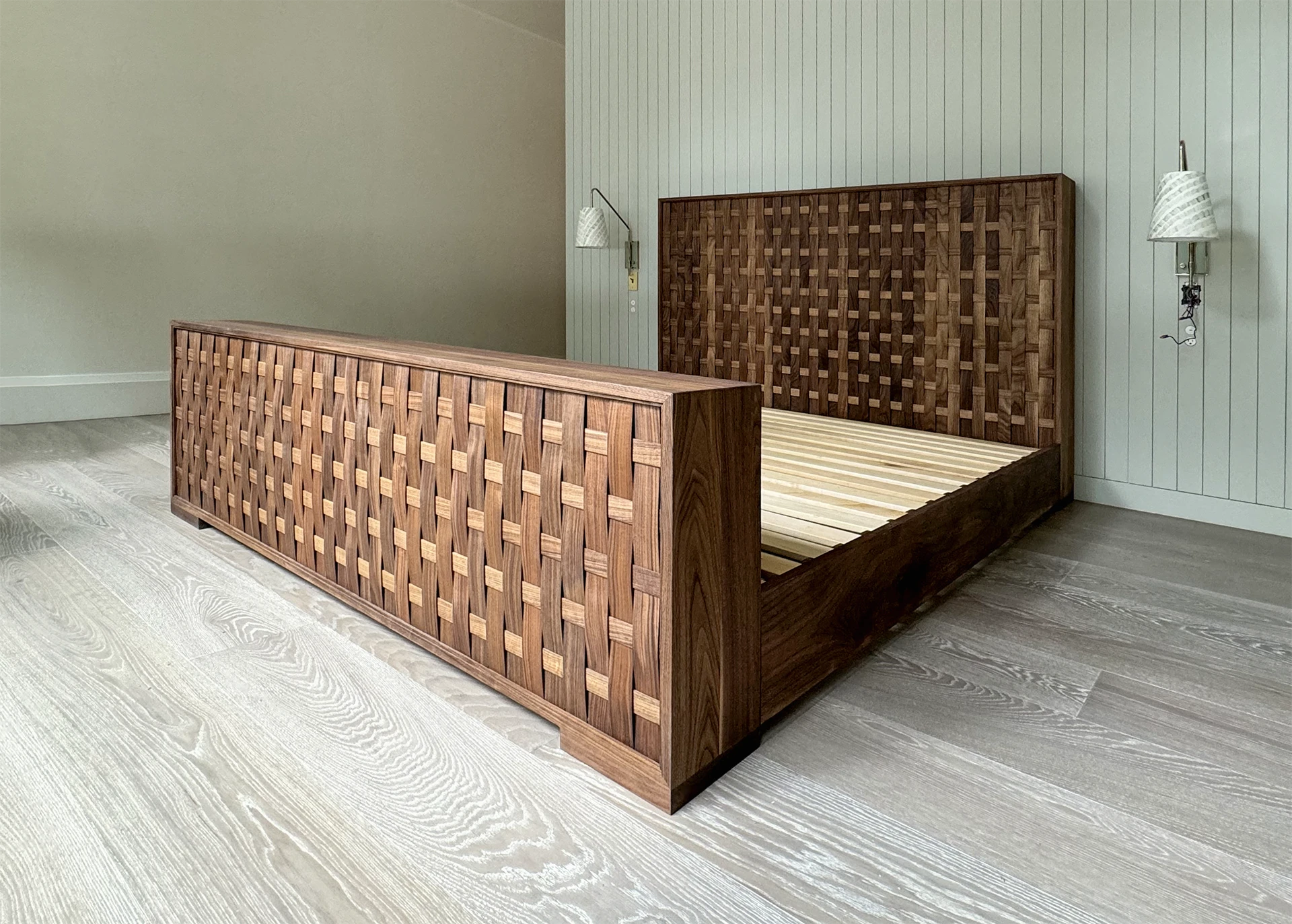 A spacious bedroom featuring a luxury walnut king size bed, with a white wood-paneled wall, grey flooring, and two white wall-mounted lamps.